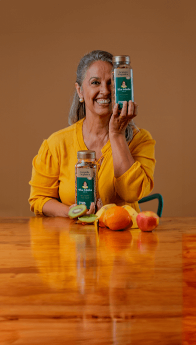 Foto com mesa de frutas e mulher segurando pote de Granola Orgânica Tia Sônia