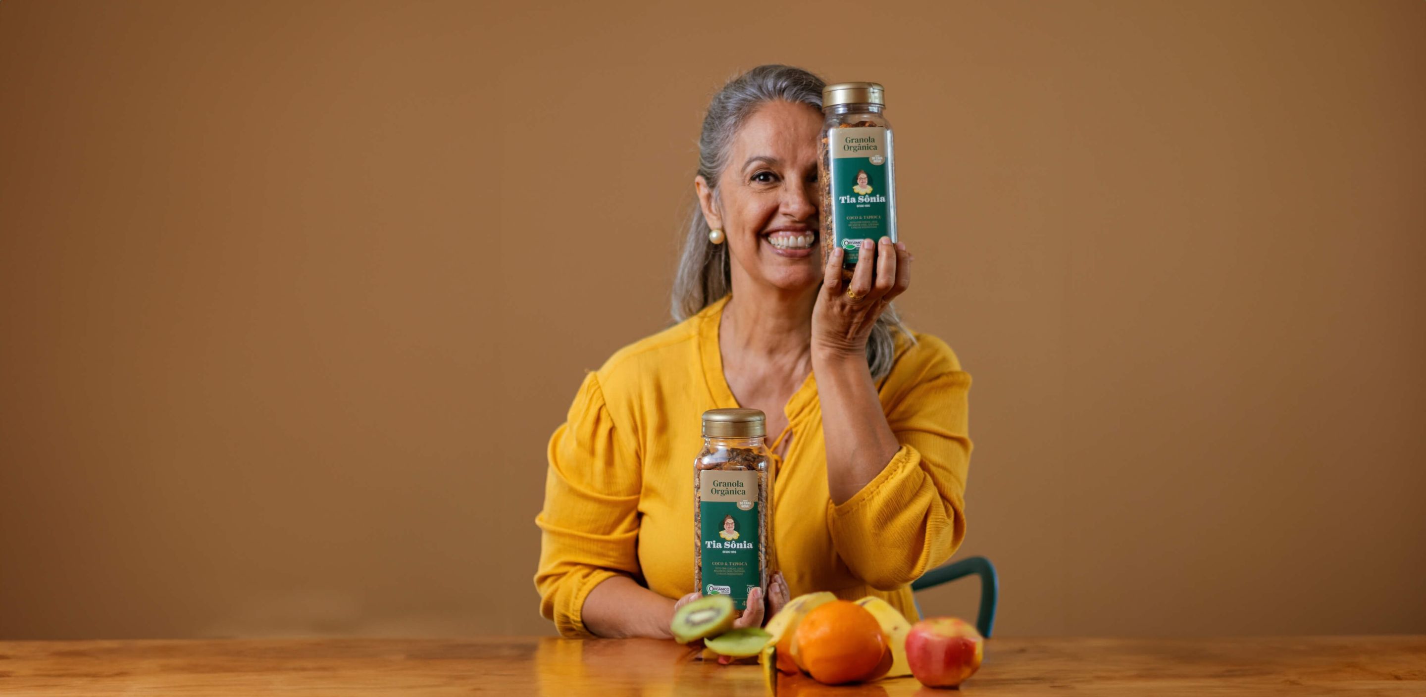 Foto com mesa de frutas e mulher segurando pote de Granola Orgânica Tia Sônia